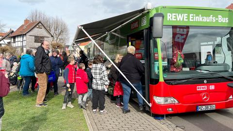 Ein Bus mit der Aufschrift "REWE-Einkaufs-Bus" an der Stirnseite steht an einer Straße. Daneben vielen Menschen.