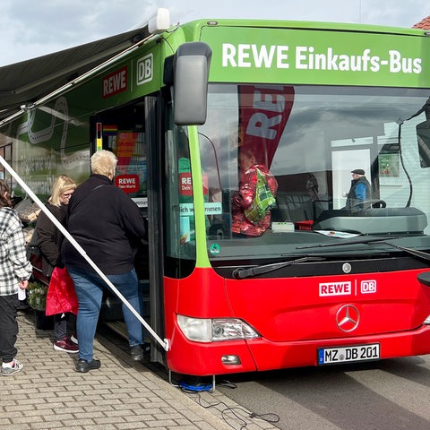 Ein Bus mit der Aufschrift "REWE-Einkaufs-Bus" an der Stirnseite steht an einer Straße. Daneben vielen Menschen.