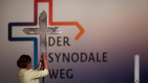 Eine Frau hält ein Kreuz hoch - dahinter steht auf einer Wand "Der Synodale Weg"