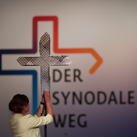 Eine Frau hält ein Kreuz hoch - dahinter steht auf einer Wand "Der Synodale Weg"