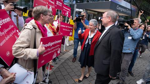 Ein Mann und eine Frau stehen einer Gruppe demonstrierender Frauen mit bunten Schildern gegenüber.