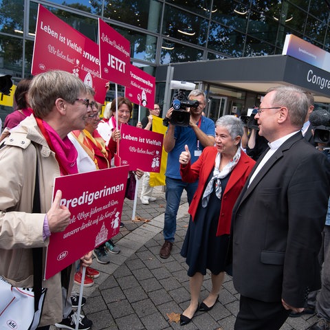 Ein Mann und eine Frau stehen einer Gruppe demonstrierender Frauen mit bunten Schildern gegenüber.