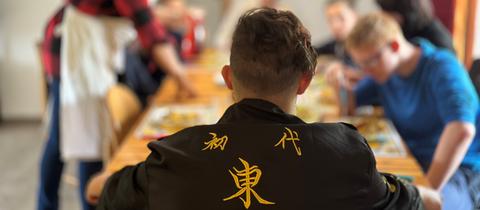 Ein Junge in einem schwarzen Oberteil mit japanischen Schriftzeichen ist von hinten zu sehen, wie er an einem Tisch mit anderen Jugendlichen sitzt