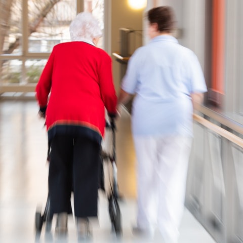 Eine Pflegerin läuft Seite an Seite mit einer älteren Bewohnerin durch den Flur eines Altenpflegeheims. (dpa)
