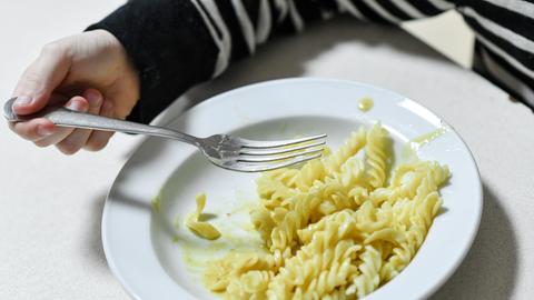 Teller mit Nudeln ohne Soße, halb leer, Kinderhand mit Gabel