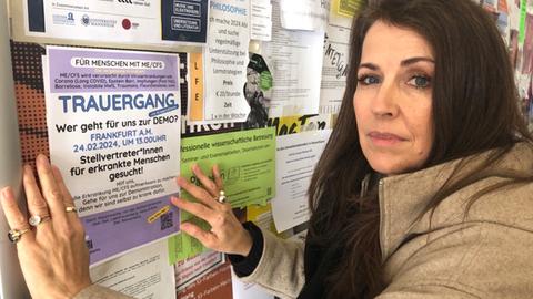 Evelyn Fay an einem Plakatbord, an dem sie ein Plakat aufhängt mit der Aufschrift: "Trauergang: Wer geht für uns zur Demo? Frankfurt 24.02.2024 um 13 Uhr - Hilf uns, auf die Erkrankung ME/CFS aufmerksam zu machen. Gehe für uns zur Demonstration, denn wir sind selbst zu krank dafür."