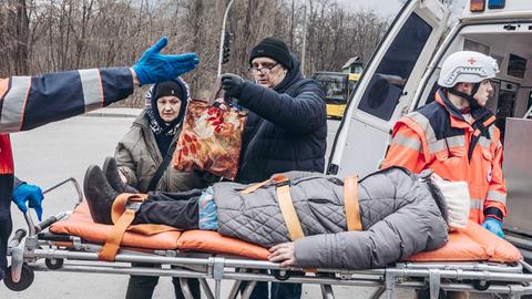Frau liegt auf einer Trage vor einem Krankenwagen. Daneben zwei Sanitäter und zwei Angehörige.