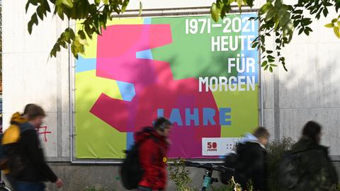 Großes Wandplakat mit der Aufschrift "1971-2021 heute für morgen, 50 Jahre", an dem jungen Menschen vorbeigehen.