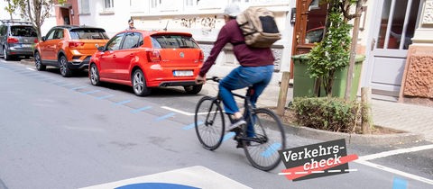Eine Straße mit parkenden Autos. In der Mitte des Bildes ein Radfahrer, leicht unscharf, weil er in Bewegung ist. Auf dem Boden der Straße eine großer blauer Kreis mit einem Fahradicon. Auf dem Bild rechts unten eine kleine Grafik mit dem Wort "Verkehrs-Check".