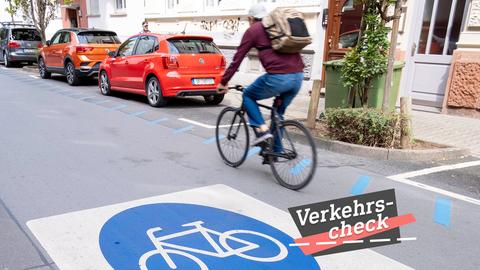 Eine Straße mit parkenden Autos. In der Mitte des Bildes ein Radfahrer, leicht unscharf, weil er in Bewegung ist. Auf dem Boden der Straße eine großer blauer Kreis mit einem Fahradicon. Auf dem Bild rechts unten eine kleine Grafik mit dem Wort "Verkehrs-Check".