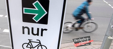 Rechts im Bild ein Radfahrer, leicht unscharf, weil er in Bewegung ist. Links davon ein Schild mit einem grünen Pfeil nach rechts, dem Wort "nur" und einem Fahradicon. Alles aus der näheren Vogelperspektive fotografiert. Auf dem Bild rechts unten eine kleine Grafik mit dem Wort "Verkehrs-Check".