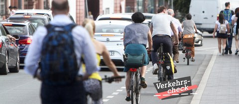 Blick eine Straße entlang, wobei mehrere Radfahrer auf einem Fahradweg zwischen Autos links und Fußgängern rechts auf dem Bürgersteig fahren. Alle sind von hinten und in Reihung zu sehen. Auf dem Bild rechts unten eine kleine Grafik mit dem Wort "Verkehrs-Check".