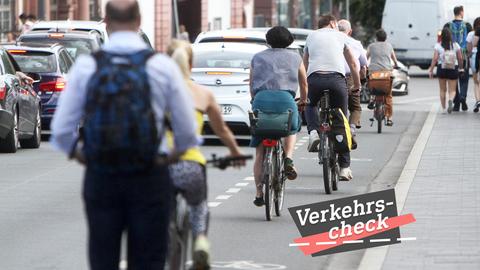 Blick eine Straße entlang, wobei mehrere Radfahrer auf einem Fahradweg zwischen Autos links und Fußgängern rechts auf dem Bürgersteig fahren. Alle sind von hinten und in Reihung zu sehen. Auf dem Bild rechts unten eine kleine Grafik mit dem Wort "Verkehrs-Check".