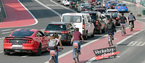 Blick eine große Straße entlang, wobei mehrere Radfahrer auf einem roten Fahradweg rechts neben Autos, die im Stau stehen., fahren. Alle sind von hinten und in Reihung zu sehen. Auf dem Bild rechts unten eine kleine Grafik mit dem Wort "Verkehrs-Check".