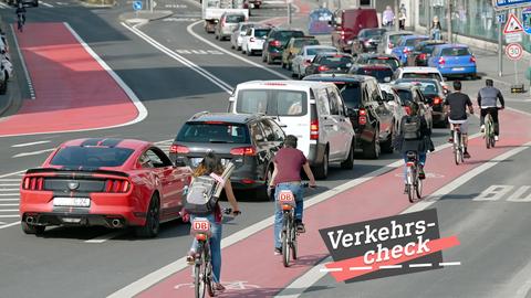 Blick eine große Straße entlang, wobei mehrere Radfahrer auf einem roten Fahradweg rechts neben Autos, die im Stau stehen., fahren. Alle sind von hinten und in Reihung zu sehen. Auf dem Bild rechts unten eine kleine Grafik mit dem Wort "Verkehrs-Check".