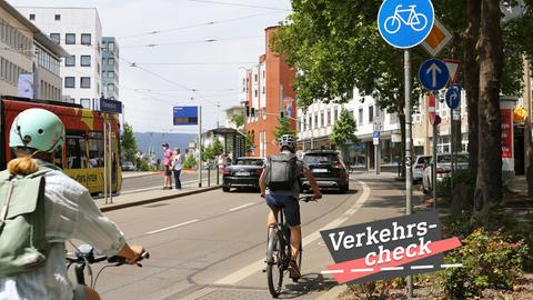 Blick in eine vierspurige Straße, daneben Radspur und Tramlinie. Radfahrer, eine Tram in Anfahrt und zwei Autos sind zu sehen. Auf dem Bild rechts unten eine kleine Grafik mit dem Wort "Verkehrs-Check".