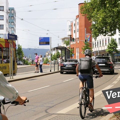 Blick in eine vierspurige Straße, daneben Radspur und Tramlinie. Radfahrer, eine Tram in Anfahrt und zwei Autos sind zu sehen. Auf dem Bild rechts unten eine kleine Grafik mit dem Wort "Verkehrs-Check".