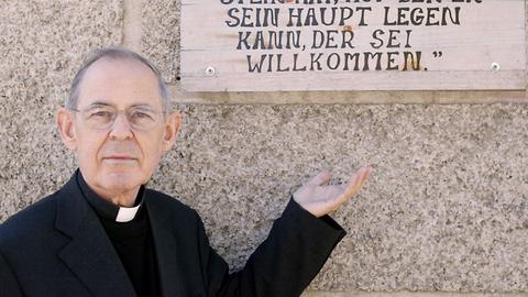 Ein älterer Pfarrer in schwarzem Anzug mit weißem Kragen zeigt auf ein Schild mit der Aufschrift "VinziDorf".