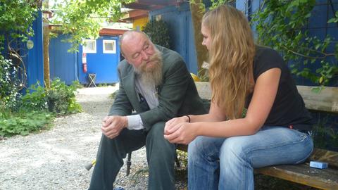 Ein älterer Mann mit Vollbart sitzt auf einer Bank und unterhält sich mit einer jungen Frau.