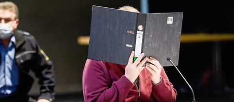 Der Angeklagte sitzt an einem Tisch mit Mikrofon in der provisorischen Gerichtshalle und hält sich einen Ordner vor das Gesicht. Er trägt eine dunkelrote Sweatshirt-Jacke.. Im Hintergrund ist ein Sicherheitsbeamter zu sehen.