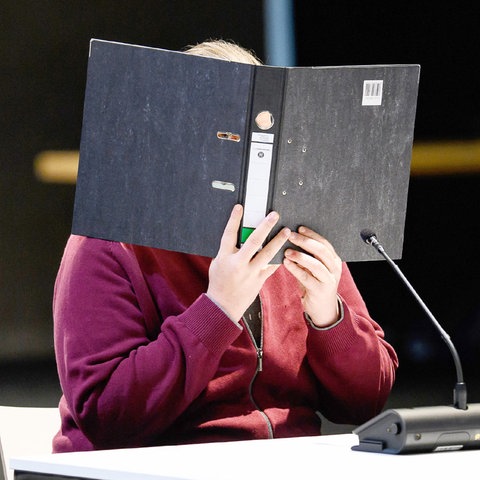 Der Angeklagte sitzt an einem Tisch mit Mikrofon in der provisorischen Gerichtshalle und hält sich einen Ordner vor das Gesicht. Er trägt eine dunkelrote Sweatshirt-Jacke.. Im Hintergrund ist ein Sicherheitsbeamter zu sehen.