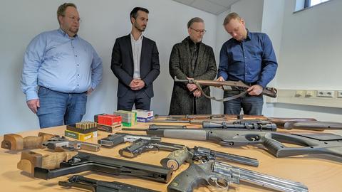 Auf einem Tisch liegen mehrere Pistolen und Gewehre. Im Hintergund stehen vier Männer. Ein Mann begutachtet ein Gewehr.