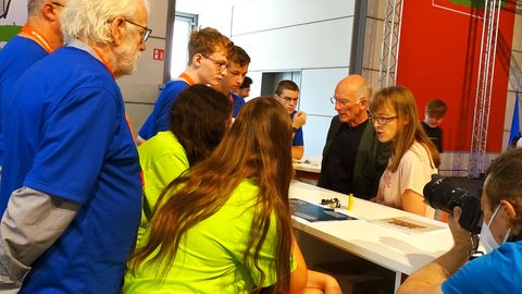 Mehrere Schülerinnen und Schüler sowie Erwachsene diskutieren an einem Messestand.