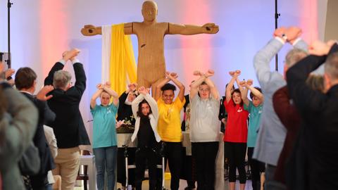 Ökumenischer Eröffungsgottesdienst der Woche für das Leben - eine Gruppe Kinder steht vor einer hölzernen Christusfigur, die die Arme ausbreitet. Die Kinder tragen dunkle Hosen und bunte T-Shirts, sie halten die Arme gekreuzt über ihren Köpfen. Einige Gottesdienstbesucher sind von hinten zu sehen. Sie machen die gleiche Handbewegung. 