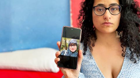 Zeinab - eine Frau mit dunklem, lockigem Haar, dunkle Brille - sie blickt ernst in die Kamera - sie hält ein Smartphone mit einem Foto, auf dem sie mit Kopftuch zu sehen ist