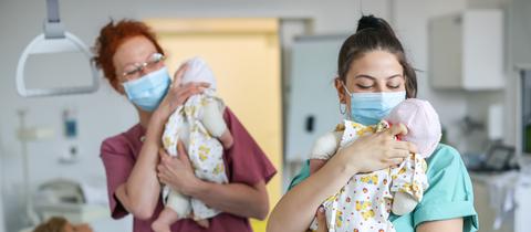 Zwei Hebammen mit Maske in einem Krankenhaus haben je einen Säugling auf dem Arm 