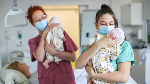 Zwei Hebammen mit Maske in einem Krankenhaus haben je einen Säugling auf dem Arm 