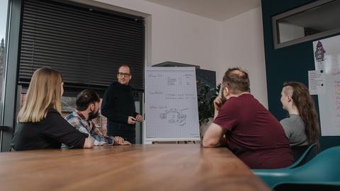 Besprechungssituation: Mehrere Menschen versammeln sich um ein Flipchart.