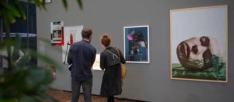 Ein Mann und eine Frau stehen vor einer Wand, an der verschiedene Fotografien hängen