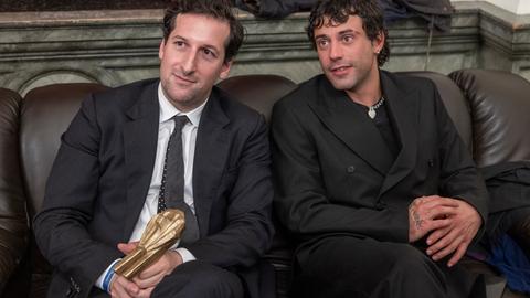 Das Bild zeigt zwei Männer, die auf dunkelbraunen Ledersitzen sitzen und schräg links aus dem Bild schauen. Sie tragen beide dunkle Anzüge. Der linke Mann hält eine goldene Auszeichnung in den Händen.