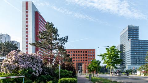 Gebäudekomplex mit rotem Klinkerbau und Hochhaus mit weiß-roter Fassade, davor Grünpflanzen