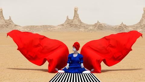 Eine sitzende Gestalt in einer Wüstenlandschaft, umweht von roten Stoffbahnen
