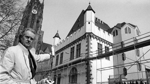 Ein Mann im Trenchcoat zeigt auf ein Haus im gotischen Stil, das von einem Baugerüst umgeben ist.