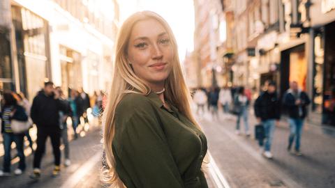 Eine junge Frau mit blonden Haaren steht in einer Straße im Gegenlicht. Es ist die Streamerin Starlet Nova. Sie trägt ein grünes, kurzes Kleid und hält eine rote Tasche in den Händen.
