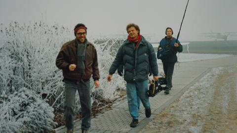 Drei Männer laufen hintereinander, sie tragen Filmequipment wie ein Mikrofon und eine Kamera.