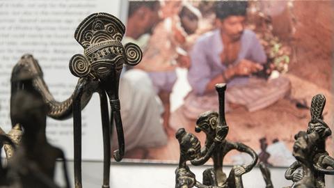 Ethnologische Objekte in einer Vitrine, im Hintergrund ein Foto von indischen Handwerkern.