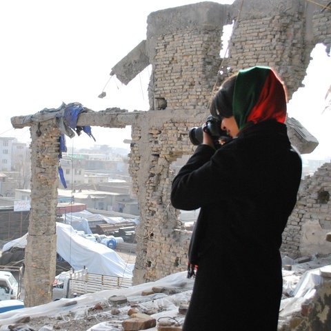 Das Bild zeigt eine Frau mit Kopftuch, die von einem erhöhten Standpunkt aus ein in Ruinen gelegtes Tal fotografiert.