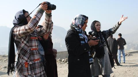 Das Bild zeigt vier Frauen mit Kopftuch, die gestikulieren und mit Spiegelreflexkameras fotografieren.