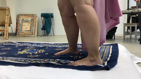 Die nackten Beine einer Frau auf einem Teppich, im Hintergrund ist ein Atelier erkennbar