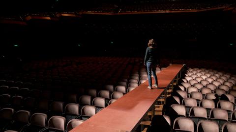 Eine Frau steht alleine auf einem Bühnensteg, der in den bestuhlten Raum ragt. Auf den Stühlen sitzt kein Publikum.