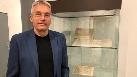 Ein Mann im blauen Sakko steht vor einer Vitrine mit antiken Notenblättern