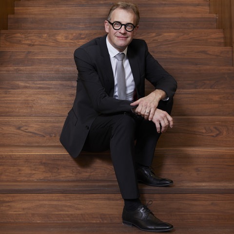 Ein Mann mit kurzen, dunkelblonden Haaren, schwarzer Hornbrille und schwarzem Anzug sitzt auf einer Holztreppe und lächelt in die Kamera.