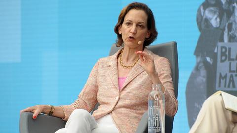 Das Bild zeigt eine Frau mit mittellangen braunen Haare und einem rosafarbenen Blazer und weißer Hose. Sie sitzt auf einem grauen Sessel auf einer Bühne und spricht in ein Mikrofon, das an ihrem Ohr befestigt ist. Ihre rechte Hand hat sie auf dem Sessel abgelegt, ihre linke auf Kinnhöhe angehoben.