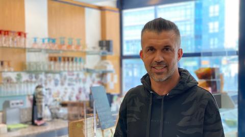 Yüksel Acun steht in seiner Eisdiele. Er trägt einen schwarzen Kapuzenpulli und einen Dreitagebart. Links von ihm sieht man verschwommen eine Eistheke und im Hintergrund Gläser in einem Regal.