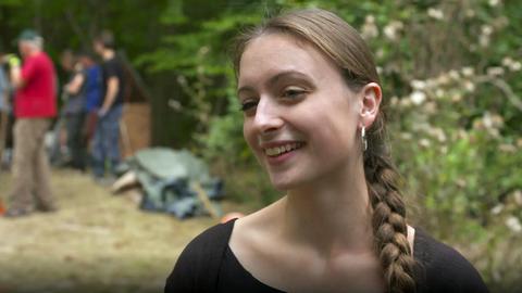 Junge Frau mit Zopf steht im Wald, im Hintergrund arbeiten Leute.