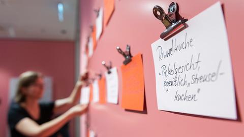 An einer rosa gestrichenen Wand hängt eine Frau weiße und orangefarbene Post-it-Zettel auf. Auf dem vordersten steht: "Riwwelkuche = Pickelgesicht, eigentlich Streuselkuchen".